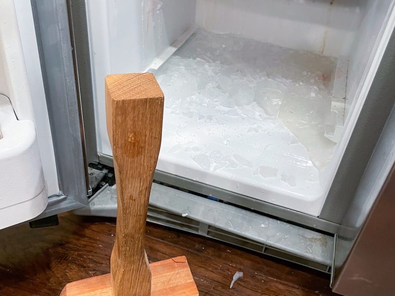 Wooden mallet in front of an open freezer with ice on the bottom.